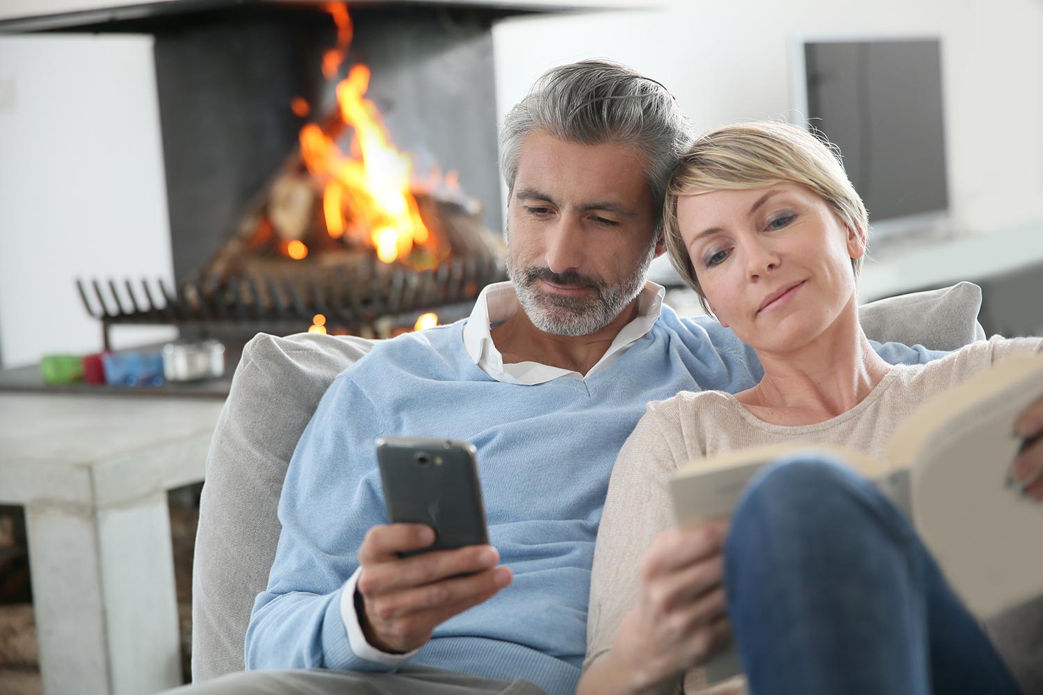 Middle-aged couple relaxing on a couch by a lit fireplace, one reading and one on a smartphone