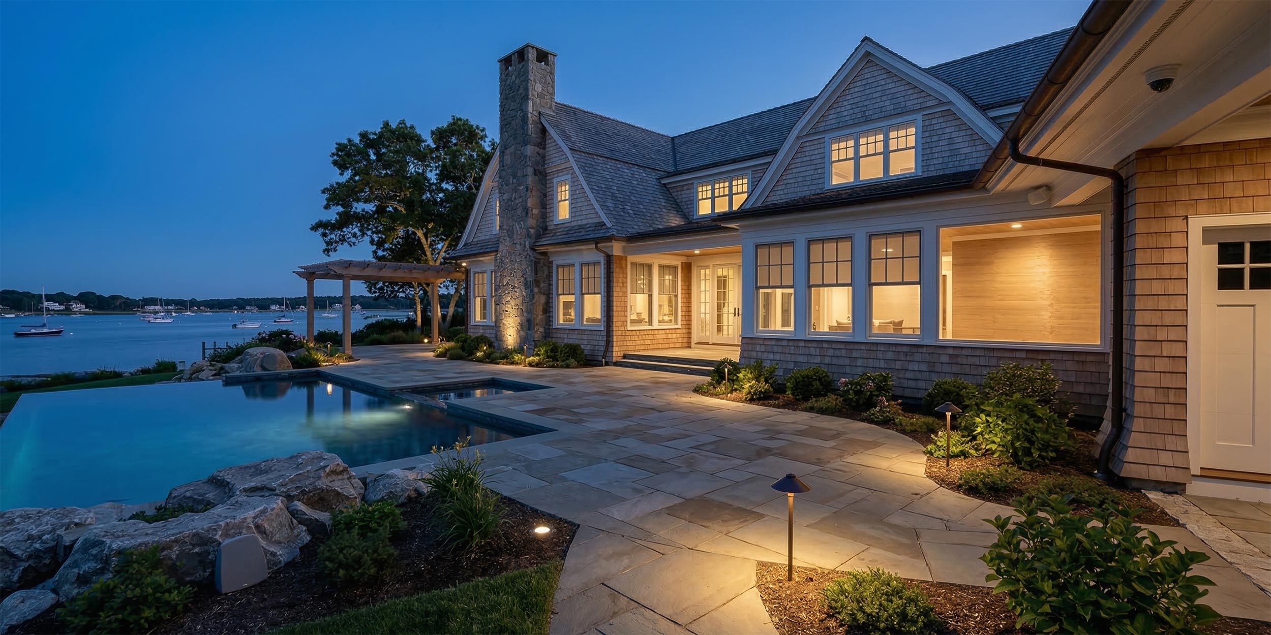 Luxury waterfront home at dusk with illuminated stone patio and infinity pool