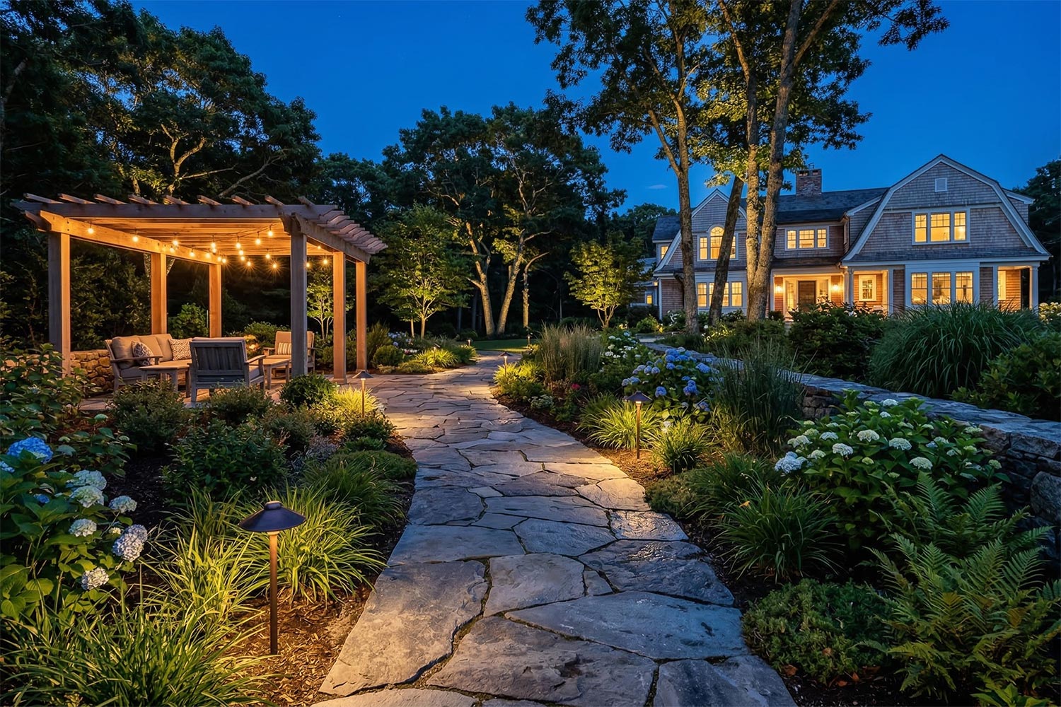 Illuminated stone pathway leading to a lit pergola and shingle-style house at night