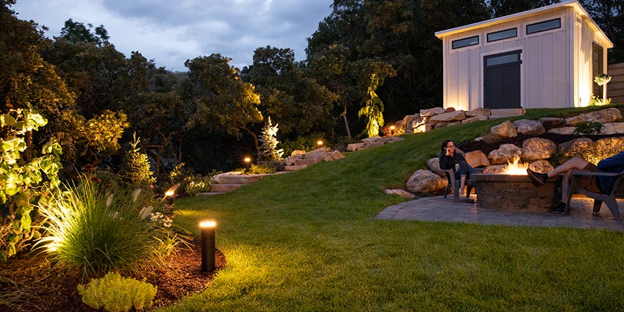 Illuminated backyard landscape at dusk with a fire pit, stone steps, and a white shed