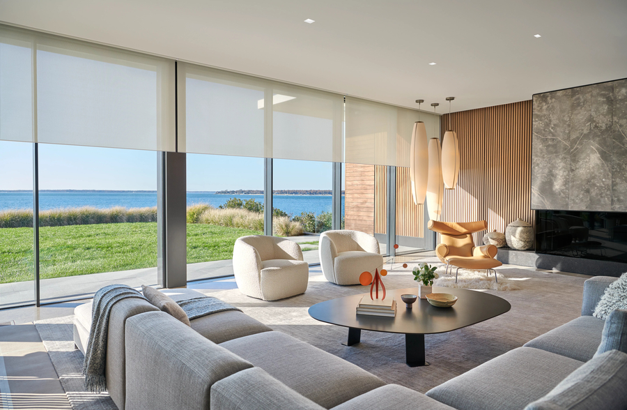 A modern, sunlit living room featuring floor-to-ceiling windows with a lake view and automated roller shades.