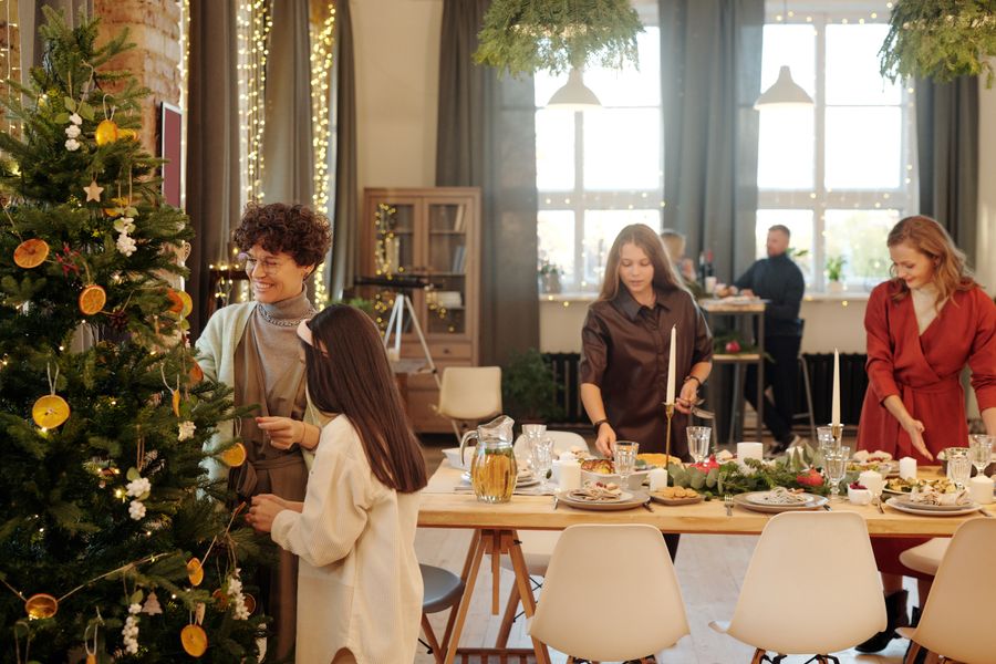<span class="uk-h4">Family and friends decorating a Christmas tree and setting a holiday dining table in a warmly lit home.</span>